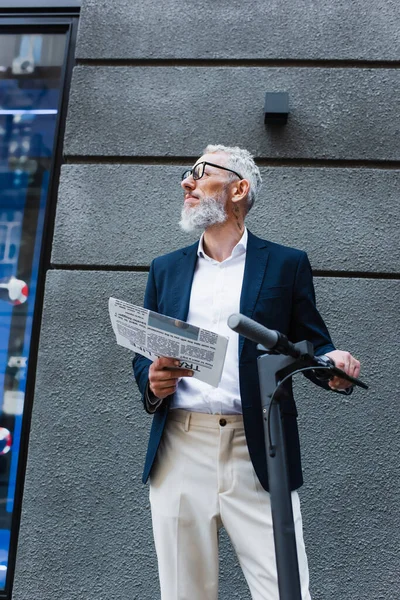 Low angle view of mature businessman in blazer holding newspaper near e-scooter - foto de stock