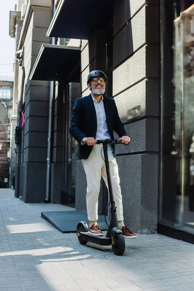 Full length of happy mature businessman in blazer and helmet riding e-scooter — Stock Photo