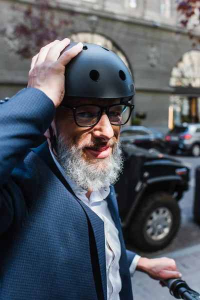 Smiling and mature man in blazer riding electric scooter and adjusting helmet - foto de stock