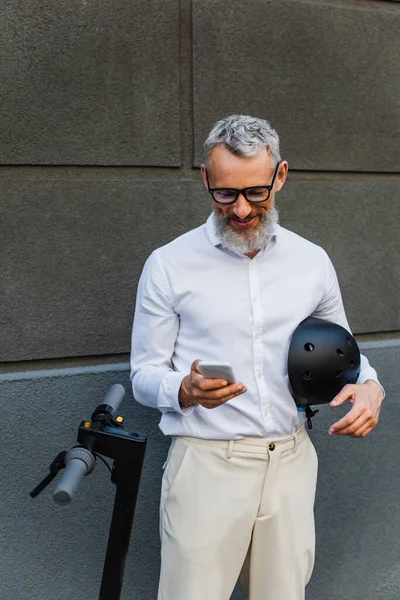 Smiling and mature man in shirt holding helmet and using smartphone near electric scooter - foto de stock