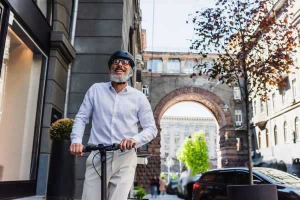 Низкоугольный вид улыбающегося взрослого мужчины в рубашке и шлеме на электрическом скутере — стоковое фото