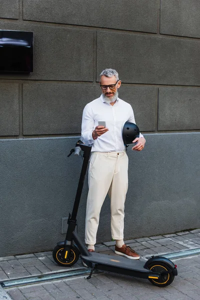 Полная длина улыбающийся и зрелый мужчина в рубашке, держа шлем и используя смартфон рядом с электрическим скутером — стоковое фото