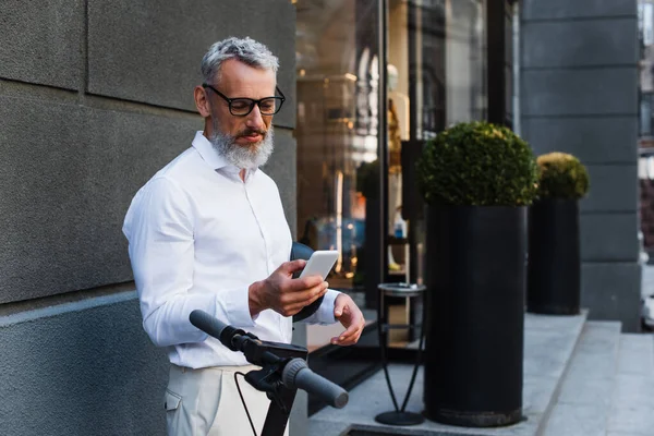 Mature man in shirt and glasses holding helmet and using smartphone near electric scooter — Foto stock