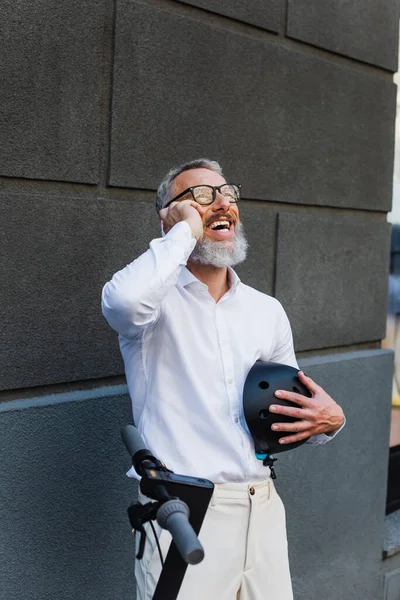 Smiling and mature man in shirt holding helmet and talking on smartphone near electric scooter - foto de stock