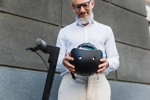 Низкий угол зрения улыбающийся и зрелый мужчина в рубашке, держащий шлем рядом с электрическим скутером — стоковое фото