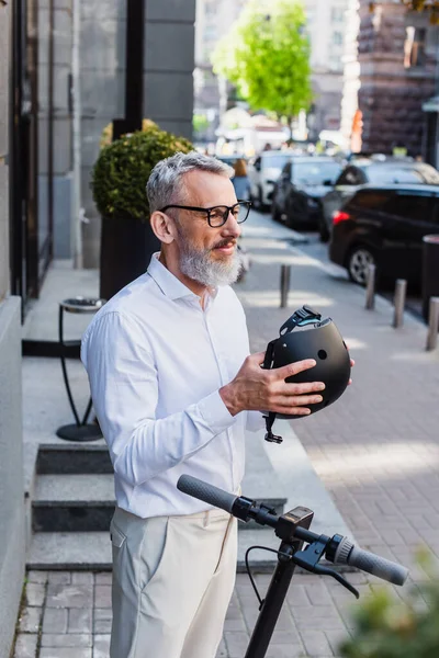 Улыбающийся и зрелый мужчина в рубашке, держащий шлем рядом с электрическим скутером — стоковое фото