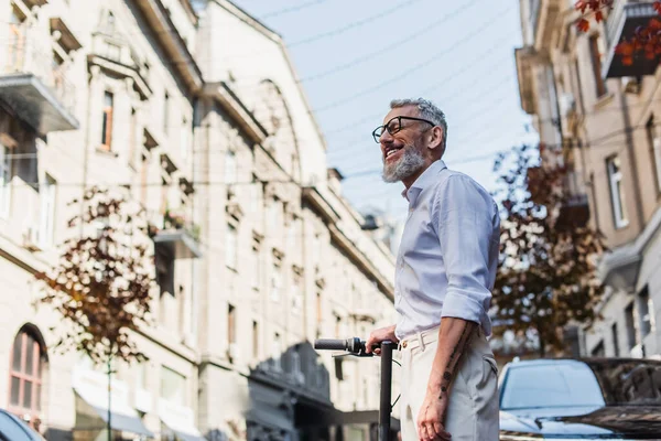 Low angle view of smiling middle aged man in white shirt near electric scooter on street - foto de stock