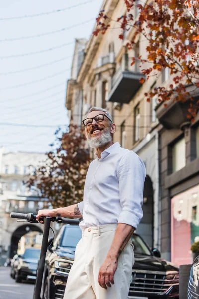 Blick auf einen fröhlichen Mann mittleren Alters in weißem Hemd, der in der Nähe eines Elektrorollers auf der Straße aufblickt — Stockfoto