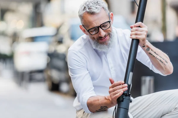 Smiling and mature man in glasses adjusting electric scooter on street — Photo de stock