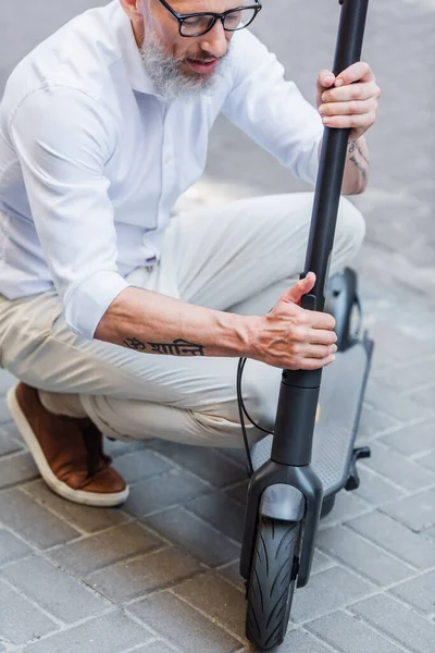 Middle aged man in glasses adjusting electric scooter on street — Stock Photo
