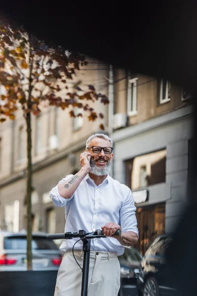 Happy middle aged man in glasses standing with electric scooter and talking on smartphone on street — Photo de stock