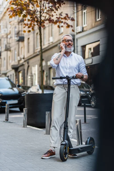 Повна довжина щасливого чоловіка середнього віку в окулярах, що розмовляють на смартфоні і стоять з електричним скутером на вулиці — стокове фото