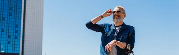 Cheerful man adjusting sunglasses near e-scooter in urban street, banner — Photo de stock