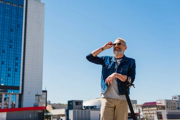 Cheerful man adjusting sunglasses near e-scooter in urban street — Stock Photo