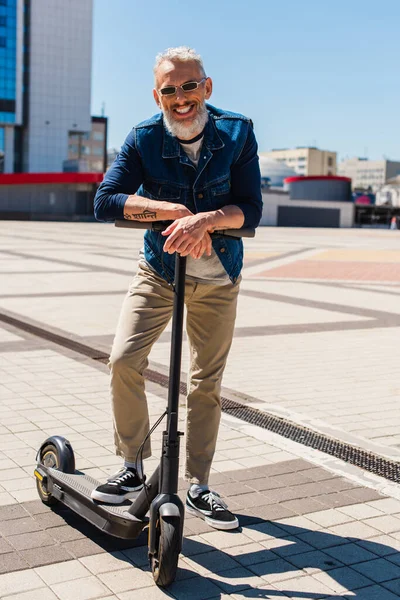 Full length of cheerful man in sunglasses standing near e-scooter in urban city — Stock Photo