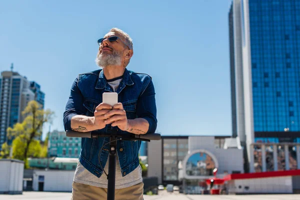 Fröhlicher Mann mit Sonnenbrille benutzt Handy in der Nähe von Elektroroller auf Stadtstraße — Stockfoto