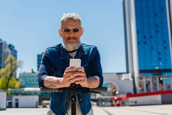 Mature man in sunglasses using cellphone near electric scooter on urban street — Photo de stock