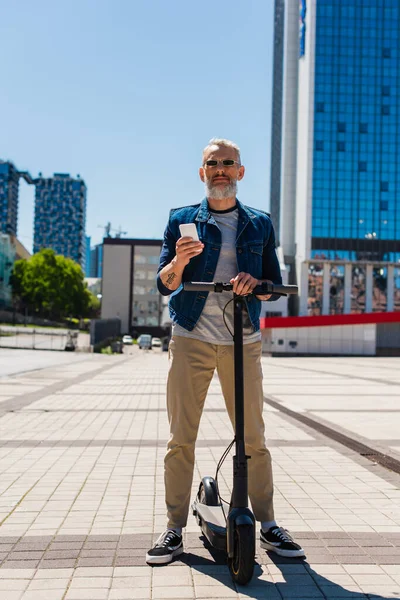Повна довжина людини в сонцезахисних окулярах, використовуючи мобільний телефон біля електричного скутера на міській вулиці — стокове фото