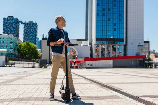 Full length of bearded man in sunglasses using cellphone near electric scooter on urban street - foto de stock
