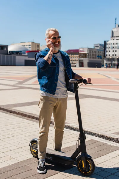 Full length of cheerful man in sunglasses talking on cellphone near electric scooter on urban street — Stock Photo