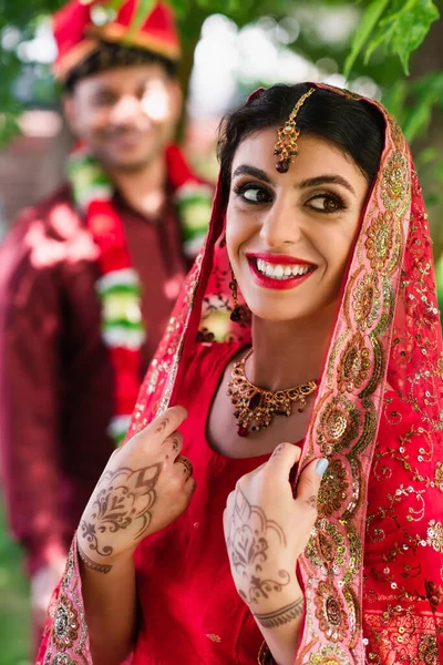 Радісна індійська наречена в сарі і хедкарф біля розмитого чоловіка в турбані на фоні — стокове фото
