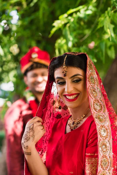 Novia india sonriente en sari y pañuelo en la cabeza cerca de hombre borroso en turbante en el fondo - foto de stock