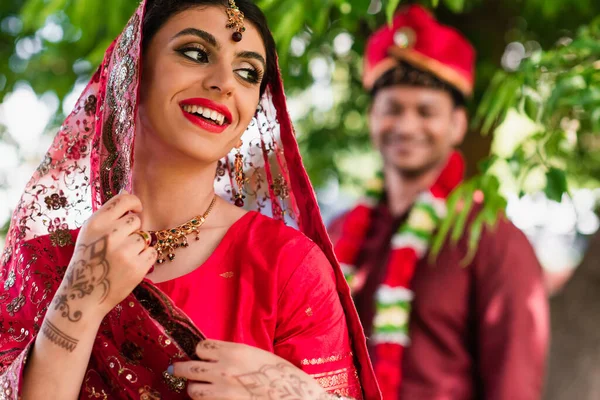 Happy indian bride in sari and headscarf near blurred man in turban on background — Stock Photo