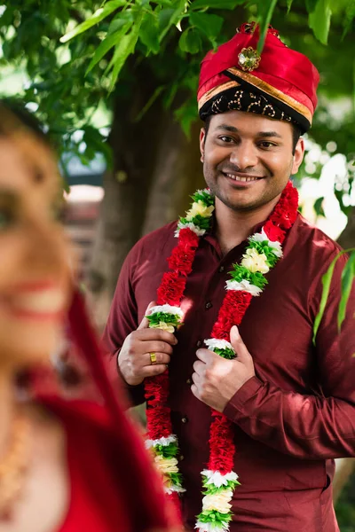 Счастливый индийский мужчина в тюрбане и цветочных гирляндах глядя на размытую невесту — стоковое фото