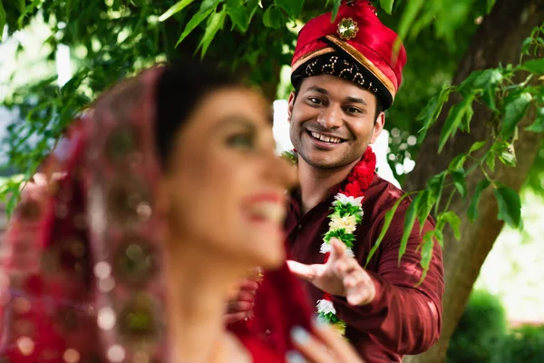 Smiling indian man in turban and floral garland reaching blurred bride — Stock Photo