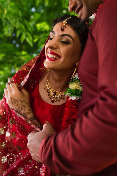 Hombre indio sonriente en turbante abrazando feliz novia en sari rojo - foto de stock