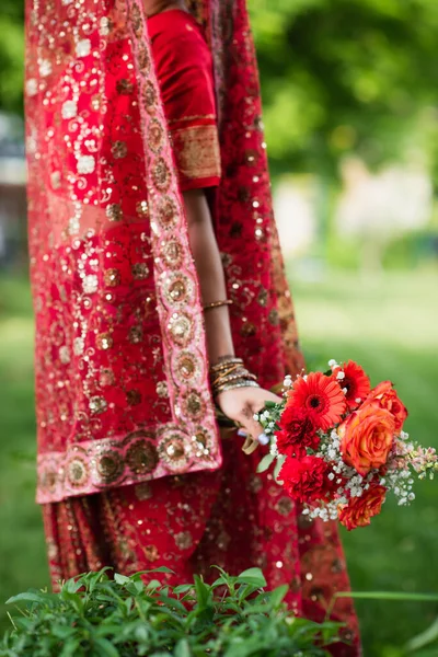 Vista cortada de noiva indiana em sari vermelho e lenço de cabeça tradicional com ornamento segurando flores — Fotografia de Stock