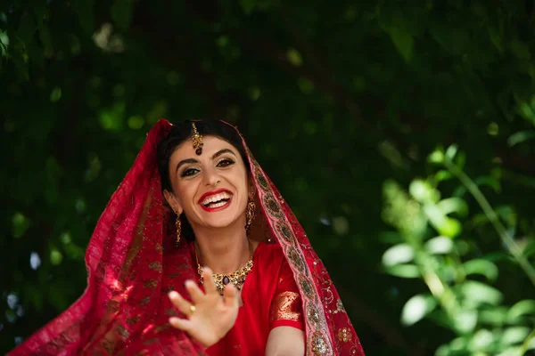 Mariée indienne heureuse en sari rouge et foulard traditionnel avec des gestes d'ornement tout en regardant la caméra — Photo de stock