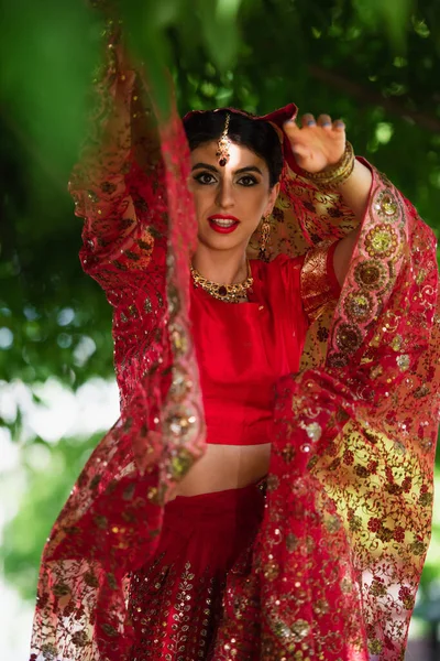 Young indian bride in red sari and traditional headscarf with ornament posing outside — Stock Photo