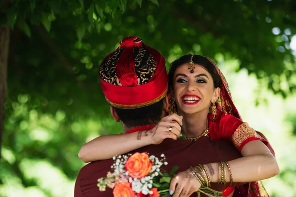 Vue arrière de l'homme indien en turban embrassant mariée joyeuse dans le foulard traditionnel tenant des fleurs — Photo de stock