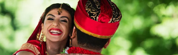 Visão traseira do homem indiano em turbante abraçando noiva feliz em lenço de cabeça tradicional, bandeira — Fotografia de Stock