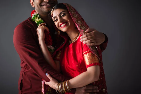Indischer Mann im Turban umarmt glückliche Braut in traditionellem Kopftuch isoliert auf grau — Stockfoto