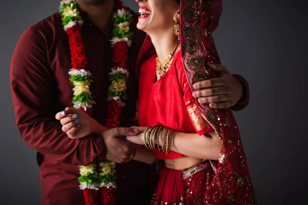 Vista recortada del hombre indio en guirnalda floral abrazando a la novia feliz en el pañuelo tradicional aislado en gris - foto de stock