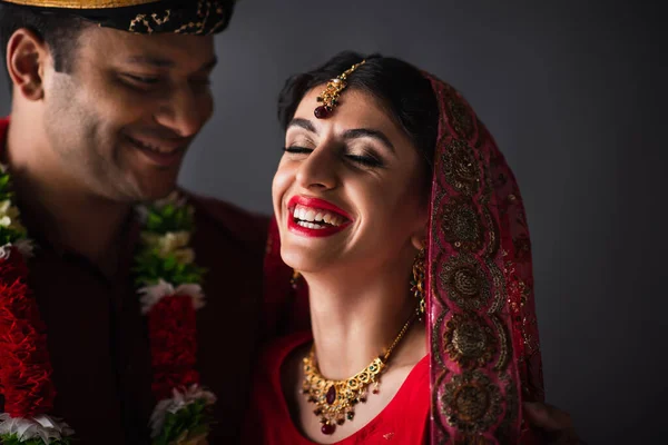 Verschwommener indischer Mann im Turban sieht glückliche Braut mit traditionellem Kopftuch isoliert auf grau an — Stockfoto