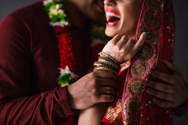Частичный вид размытого индийского мужчины в тюрбане, держащего за руку счастливую невесту в традиционном платке, изолированном на сером — стоковое фото