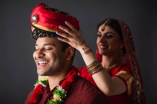 Cheerful indian bride wearing turban on head of bridegroom isolated on grey — Stock Photo