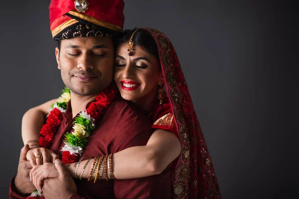 Fröhliche indische Braut im Sari umarmt Bräutigam im Turban isoliert auf grau — Stockfoto