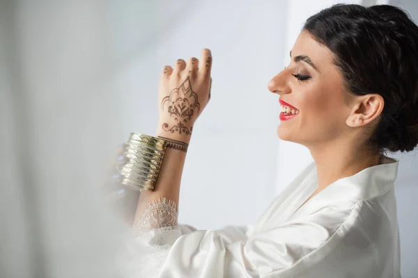 Young and happy indian bride smiling and wearing golden bracelet on white — Stock Photo