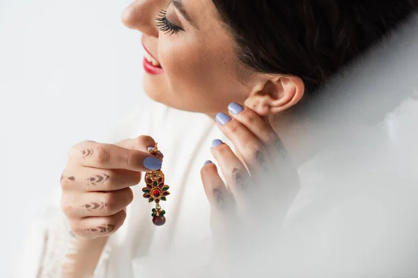 Smiling indian bride wearing earring while getting ready for wedding isolated on white — Stock Photo