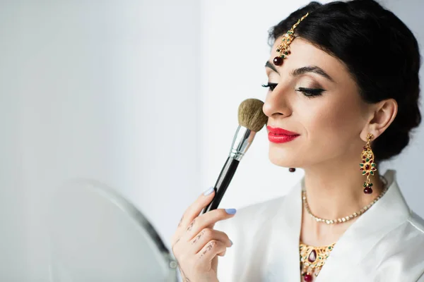 Indian bride with mehndi applying face powder with cosmetic brush near mirror on white — Stock Photo