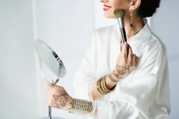Vue recadrée de mariée indienne heureuse avec mehndi appliquer la poudre visage avec pinceau cosmétique sur blanc — Photo de stock