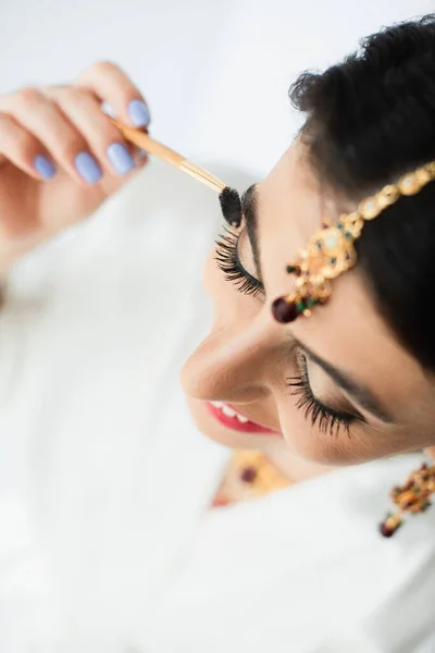 Top view of happy indian bride with mehndi styling eyebrow with cosmetic brush on white — Stock Photo