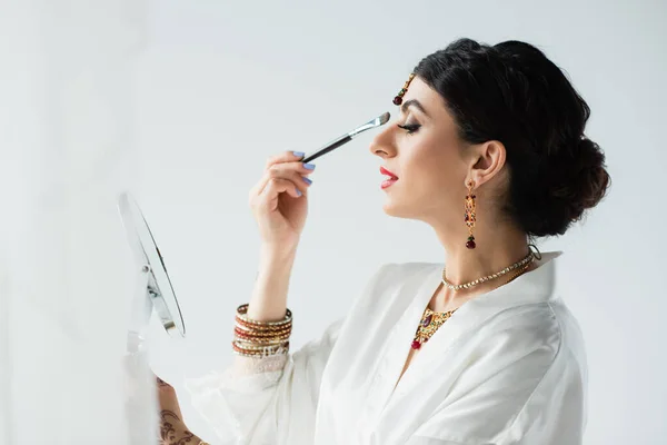 Side view of indian bride with mehndi applying eye shadow with cosmetic brush on white — Stock Photo