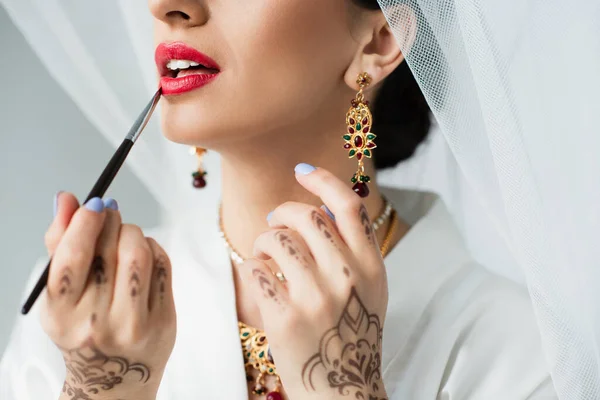 Vista recortada de la novia india aplicando lápiz labial con cepillo cosmético sobre blanco - foto de stock