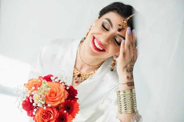 Jovem noiva indiana alegre com mehndi segurando buquê de flores no branco — Fotografia de Stock