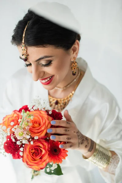 Jeune mariée indienne joyeuse avec mehndi regardant bouquet de fleurs sur blanc — Photo de stock